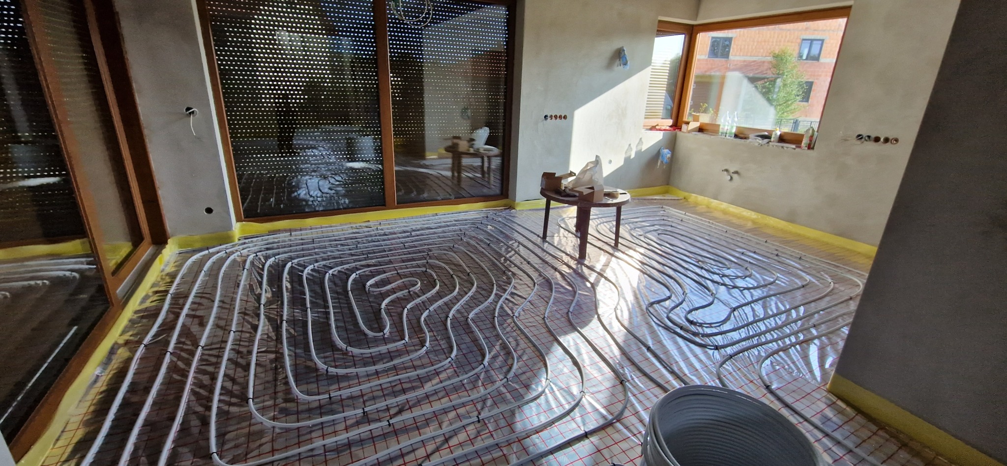 Białe rury ogrzewania podłogowego ułożone na folii izolacyjnej z siatką, widoczne w surowym wnętrzu domu w trakcie instalacji, z oknem w tle i stolikiem z narzędziami.