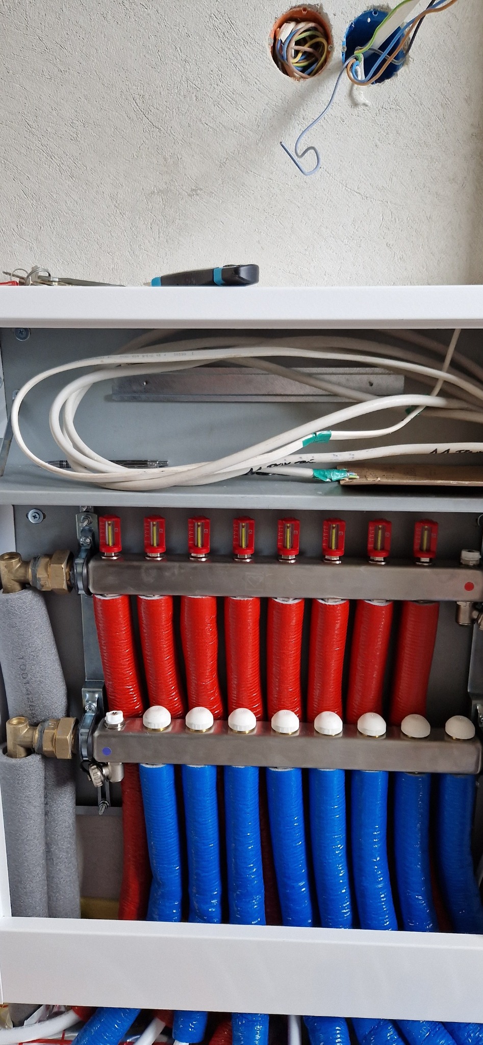 Rozdzielacz ogrzewania podłogowego z czerwonymi i niebieskimi rurami, widoczne okablowanie elektryczne w ścianie i narzędzia na półce powyżej.
