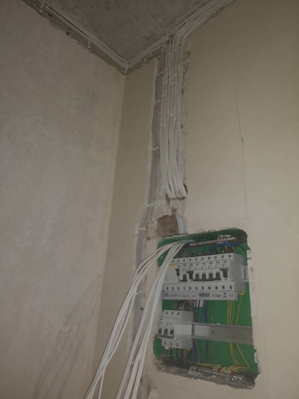 Ściana w surowym stanie z widocznymi przewodami elektrycznymi prowadzącymi do skrzynki rozdzielczej z zielonym obramowaniem, widoczne zabezpieczenia i kable wewnątrz skrzynki.