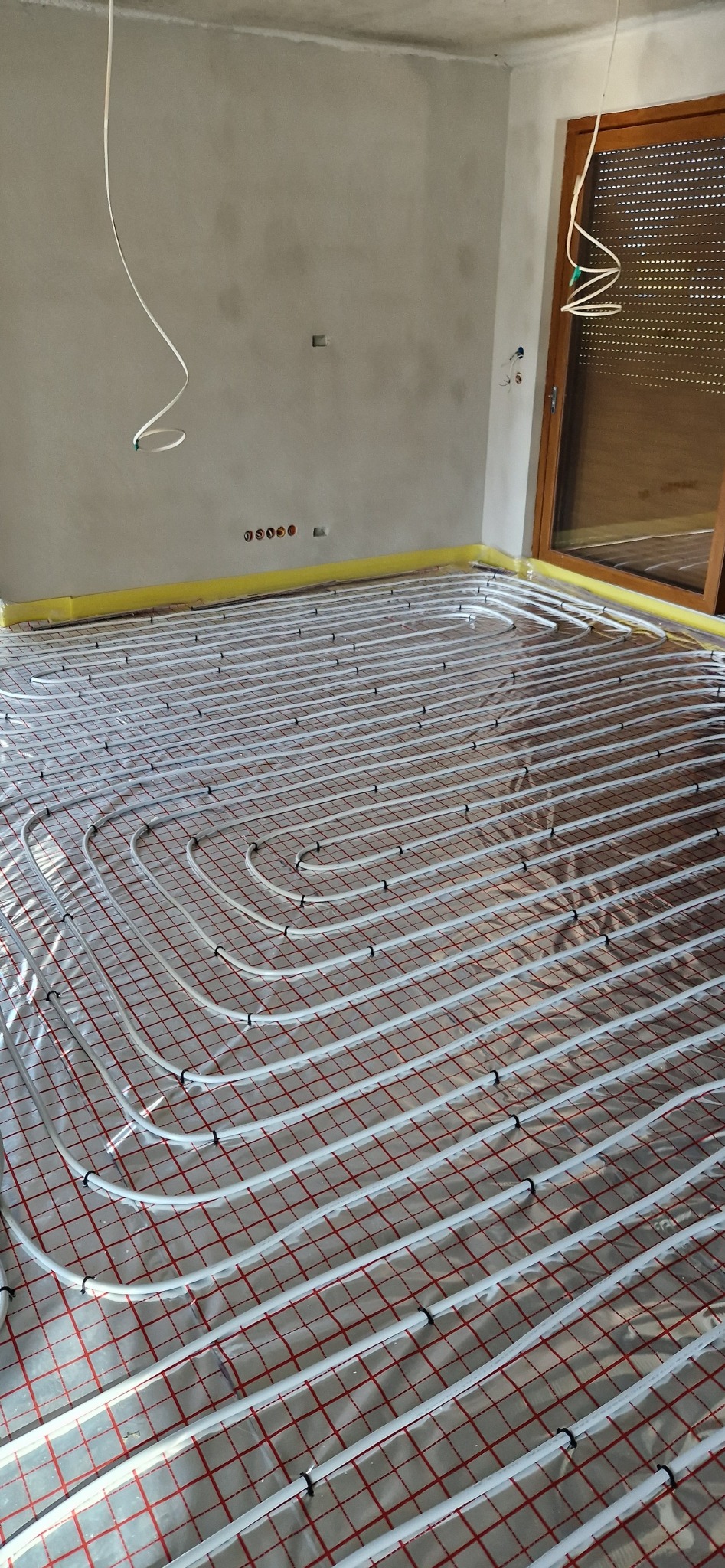 Białe rury ogrzewania podłogowego ułożone na czerwonej folii w surowym wnętrzu z szarymi ścianami i żółtą taśmą izolacyjną wzdłuż krawędzi.