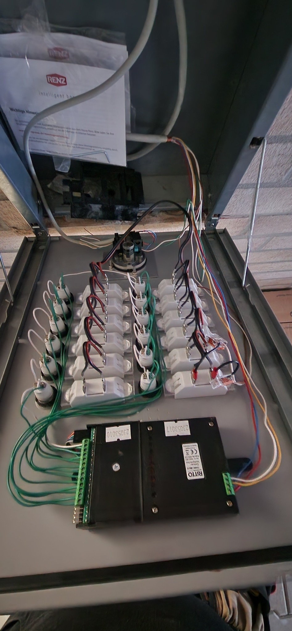 Otwarta skrzynka rozdzielcza z licznymi przewodami i złączkami, widoczne dwa czarne moduły RITO z zielonymi listwami zaciskowymi, dokumentacja Renz w tle.