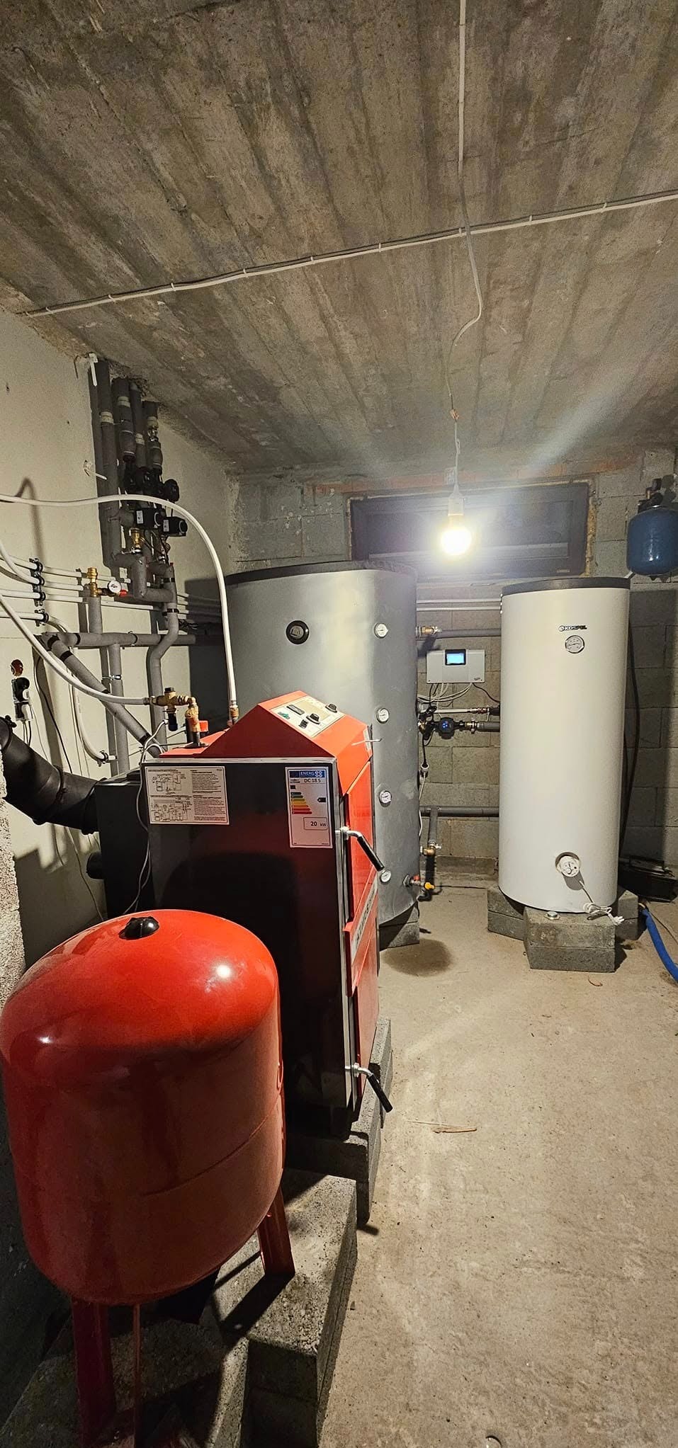 Kotłownia w piwnicy z kotłem na paliwo stałe, zbiornikiem akumulacyjnym i zasobnikiem ciepłej wody użytkowej, widoczne rury instalacji grzewczej i oświetlenie żarówką