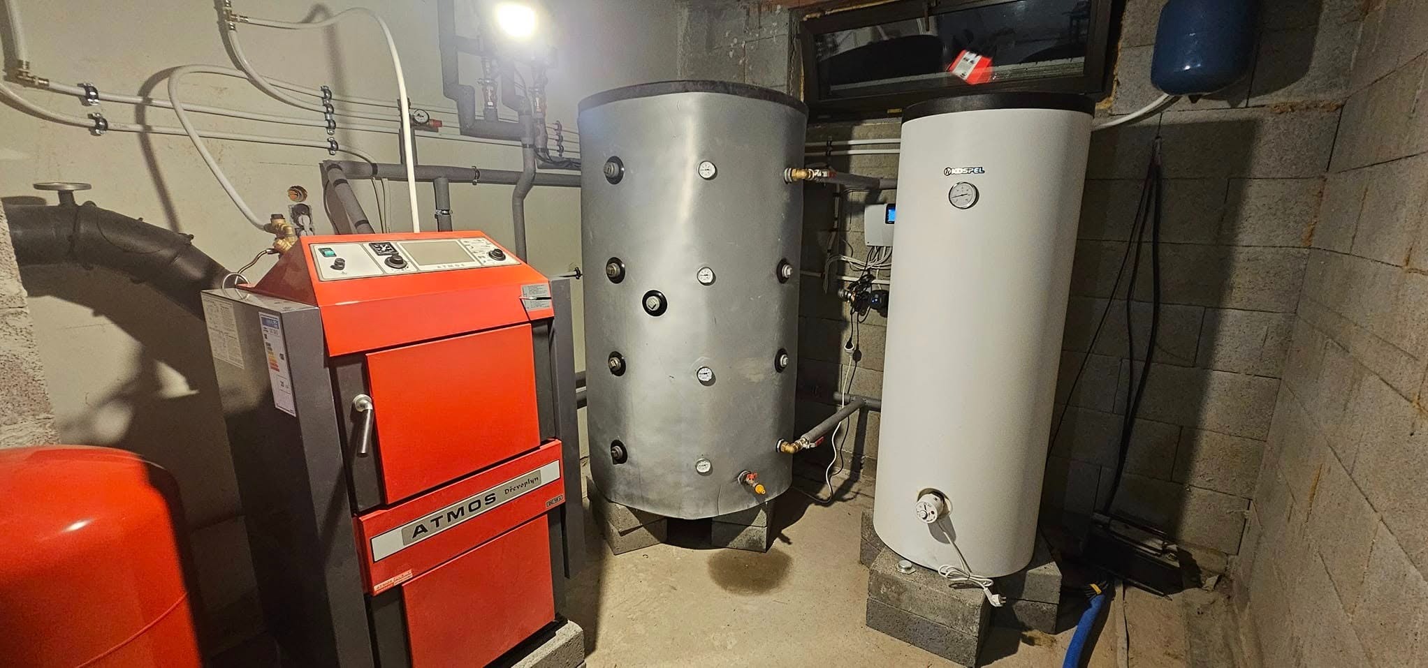 Kotłownia z piecem Atmos na paliwo stałe, dwoma zbiornikami akumulacyjnymi na betonowych podstawach i instalacją rurową przy ścianie z bloków betonowych.