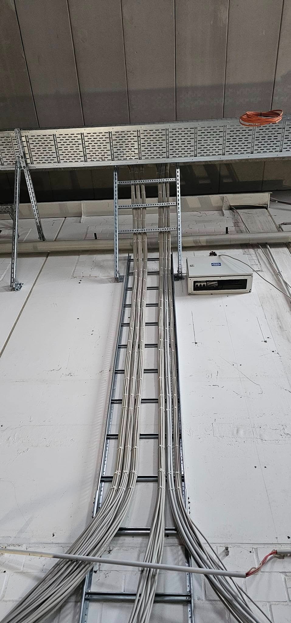 Szczegółowe ujęcie pionowego koryta kablowego z licznymi przewodami elektrycznymi, zamocowanego do ściany z widoczną fakturą, z dodatkowym korytem pod sufitem i zwiniętym kablem na nim.