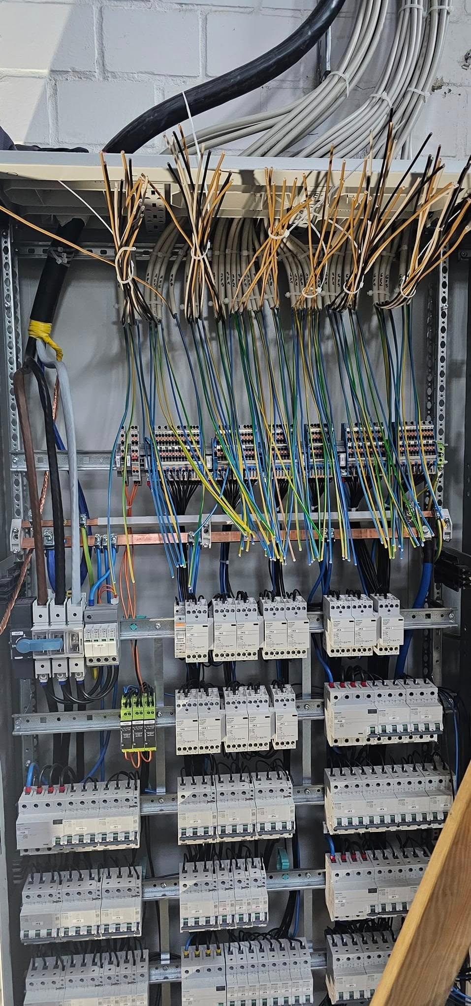 Otwarta szafa elektryczna z widocznymi przewodami o różnych kolorach (niebieski, żółty, zielony, czarny) i zabezpieczeniami, w tle szara ściana z cegieł.