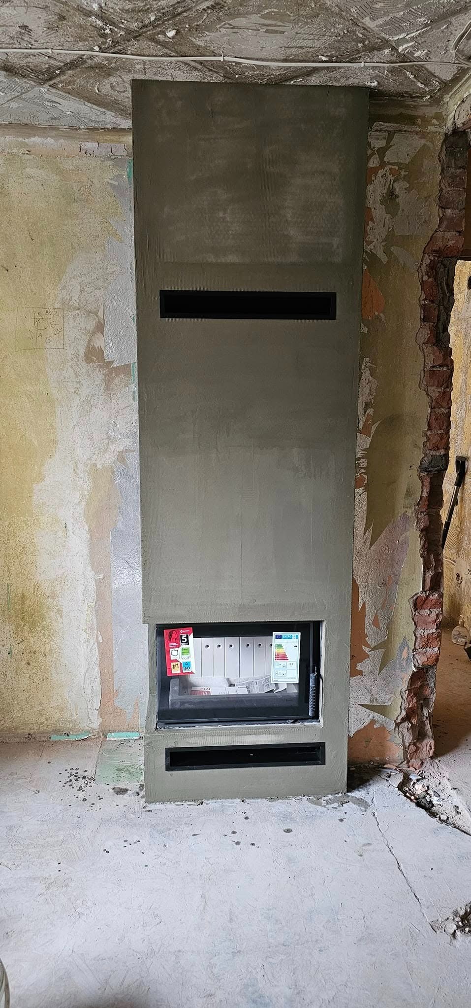 Surowy kominek w trakcie wykończenia, z widocznymi elementami instalacji i fragmentami ceglanej ściany w tle.