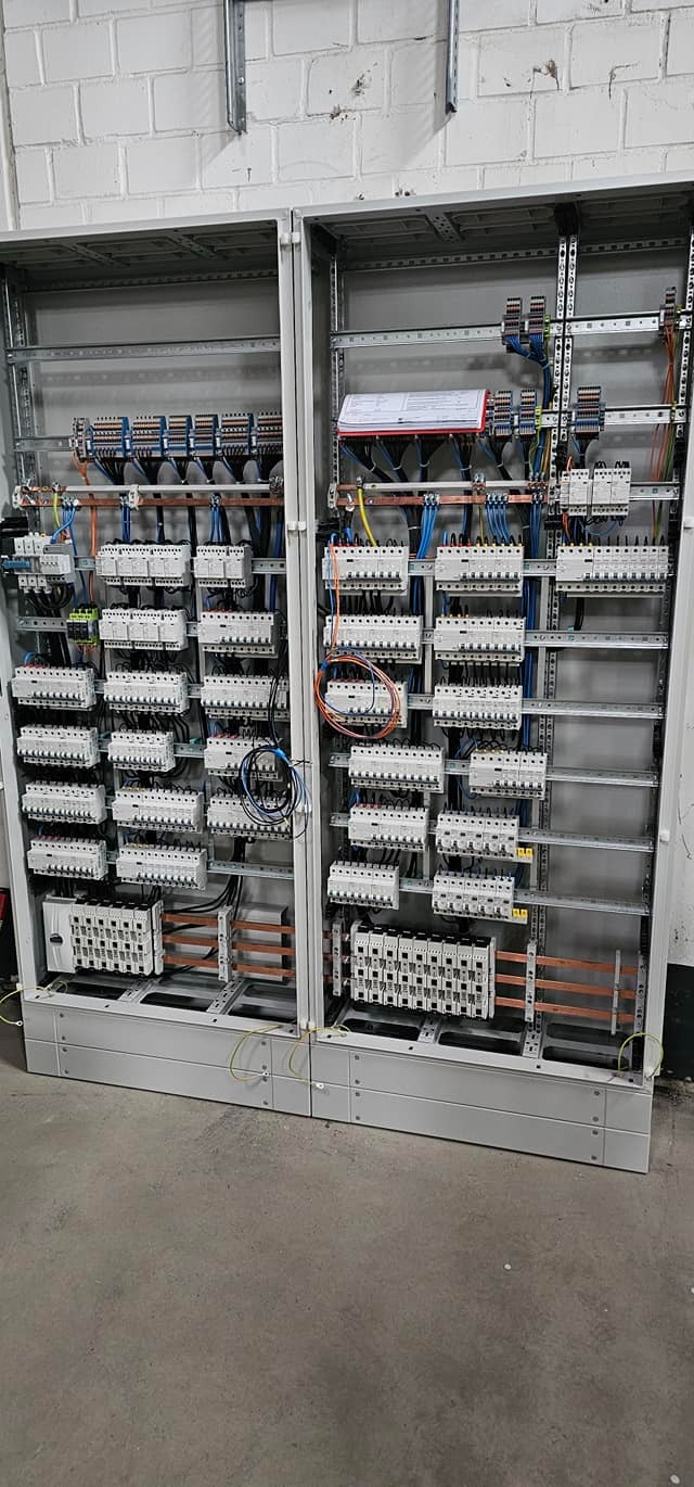 Dwie szafy elektryczne w trakcie montażu, widoczne liczne zabezpieczenia, przewody i listwy zaciskowe, na górze jednej z szaf dokumentacja techniczna.