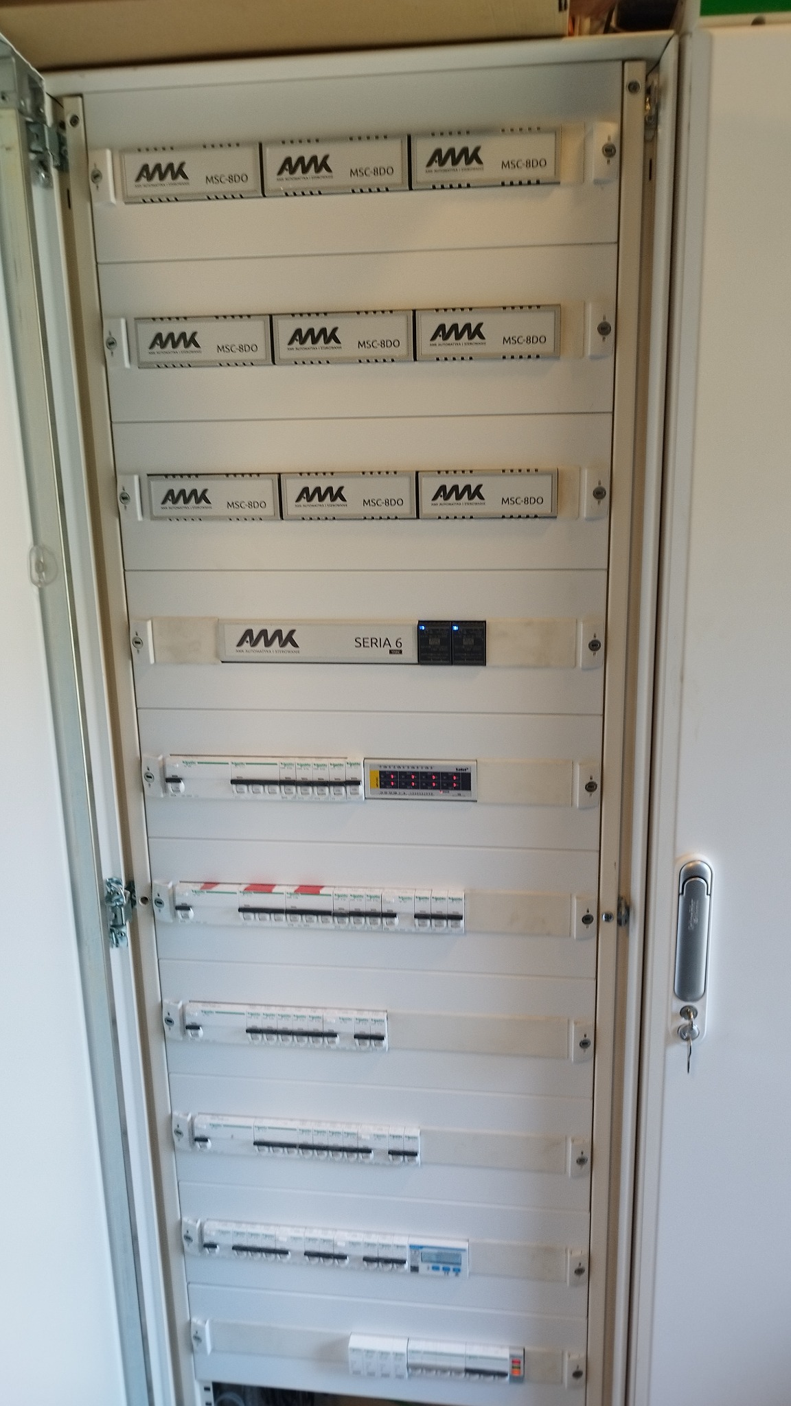 Otwarta, biała szafa elektryczna z zamontowanymi modułami AMK MSC-8DO i bezpiecznikami, widoczne liczne zabezpieczenia i wyłączniki różnicowoprądowe.