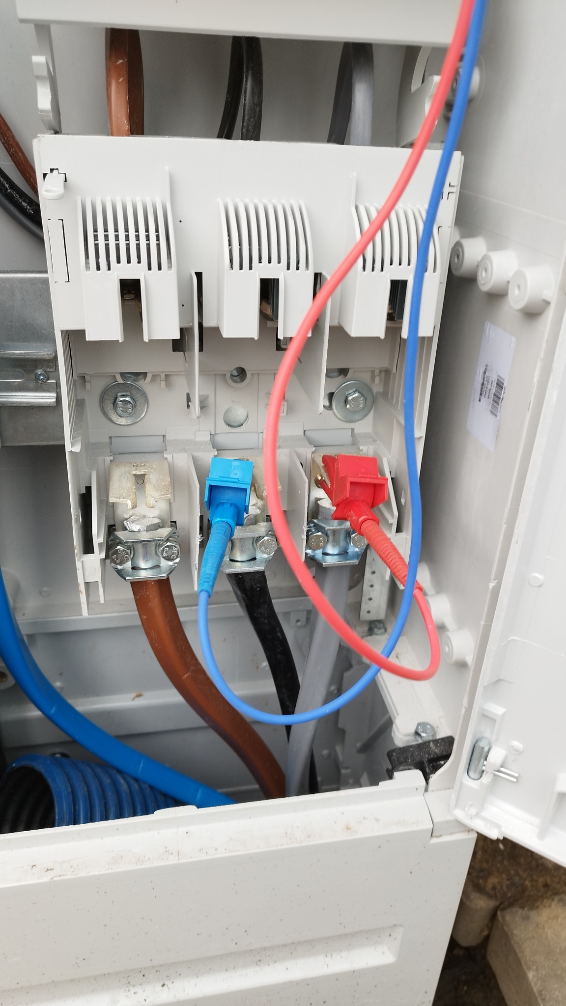 Otwarta skrzynka elektryczna z podłączonymi przewodami o różnych kolorach (niebieski, czerwony, brązowy, czarny, szary) do zacisków śrubowych, widoczne elementy montażowe i fragmenty obudowy.