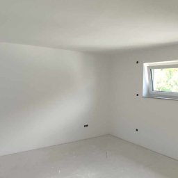 Wall Done Olaf Karbowniczek - Wnętrze pokoju po nałożeniu białej warstwy wykończeniowej na ściany i podłogę, z widocznym oknem i fragmentem surowej ściany z cegły i betonu.