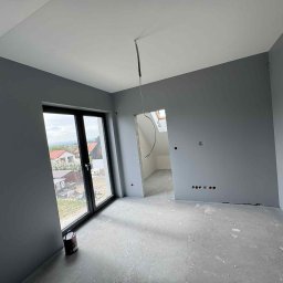 Wall Done Olaf Karbowniczek - Pomieszczenie po malowaniu na szaro, widoczne okno z widokiem na domy, puszka farby przy ścianie, surowy stan wykończenia przy otworze drzwiowym.