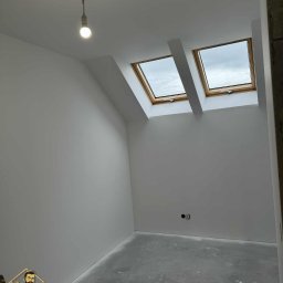 Wall Done Olaf Karbowniczek - Jasne poddasze po remoncie z dwoma oknami dachowymi i szarą podłogą. Widoczna żarówka na kablu i logo firmy w rogu. Ściany świeżo pomalowane na biało.