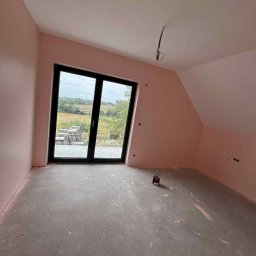 Wall Done Olaf Karbowniczek - Pokój na poddaszu po malowaniu ścian na różowo, widoczne okno z widokiem na zewnątrz, surowa podłoga i puszka farby. Ściany pomalowane na jednolity kolor.