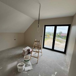 Wall Done Olaf Karbowniczek - Wykończone ściany w odcieniach beżu w pokoju na poddaszu z oknem, drabina, worki z gładzią i puszki z farbą na podłodze. Widok na zewnątrz przez okno.