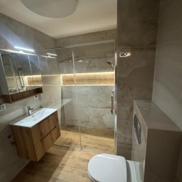 Wall Done Olaf Karbowniczek - Nowoczesna łazienka z kabiną prysznicową typu walk-in, drewnianą szafką pod umywalką i beżowymi płytkami imitującymi marmur. Oświetlenie LED podkreśla detale.