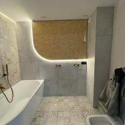 Wall Done Olaf Karbowniczek - Wykończona łazienka z mozaikową podłogą i ścianą, podświetlona LED. Widoczna wanna, toaleta i fragment ściany z surową płytą OSB. Armatura w kolorze miedzi.