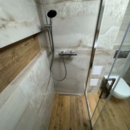 Wall Done Olaf Karbowniczek - Nowoczesna kabina prysznicowa typu walk-in z drewnopodobną podłogą i marmurowymi płytkami ściennymi, widoczny odpływ liniowy i toaleta w tle.