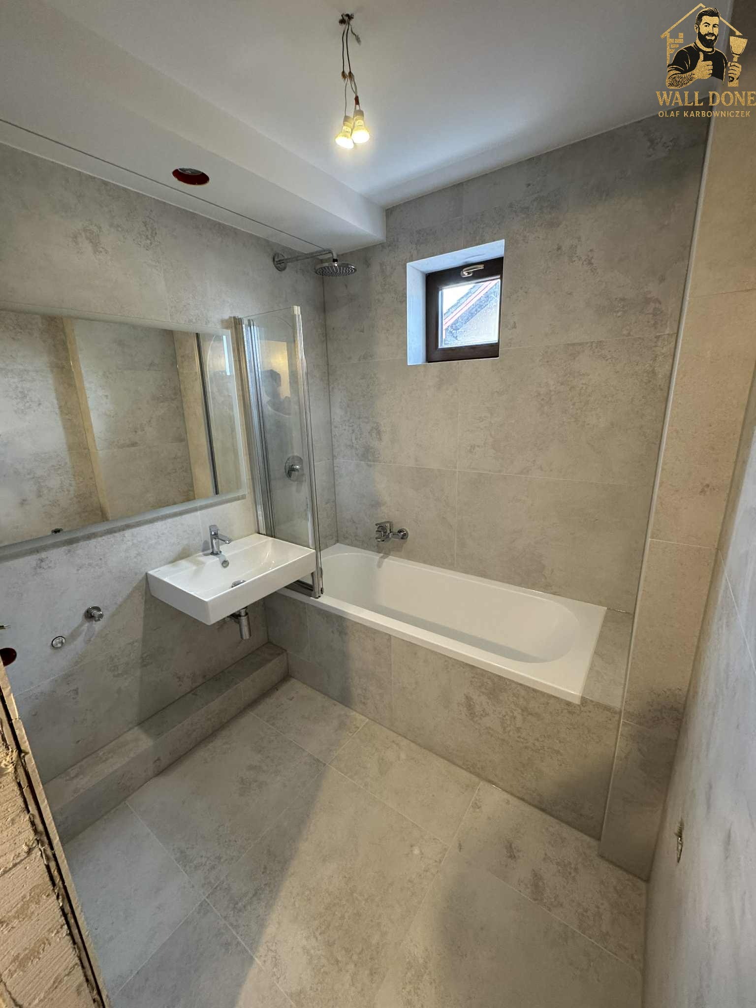Jasna łazienka po remoncie z wanną, umywalką i szklanym parawanem. Szare płytki na ścianach i podłodze. Małe okno z drewnianą ramą. Widoczne logo firmy.
