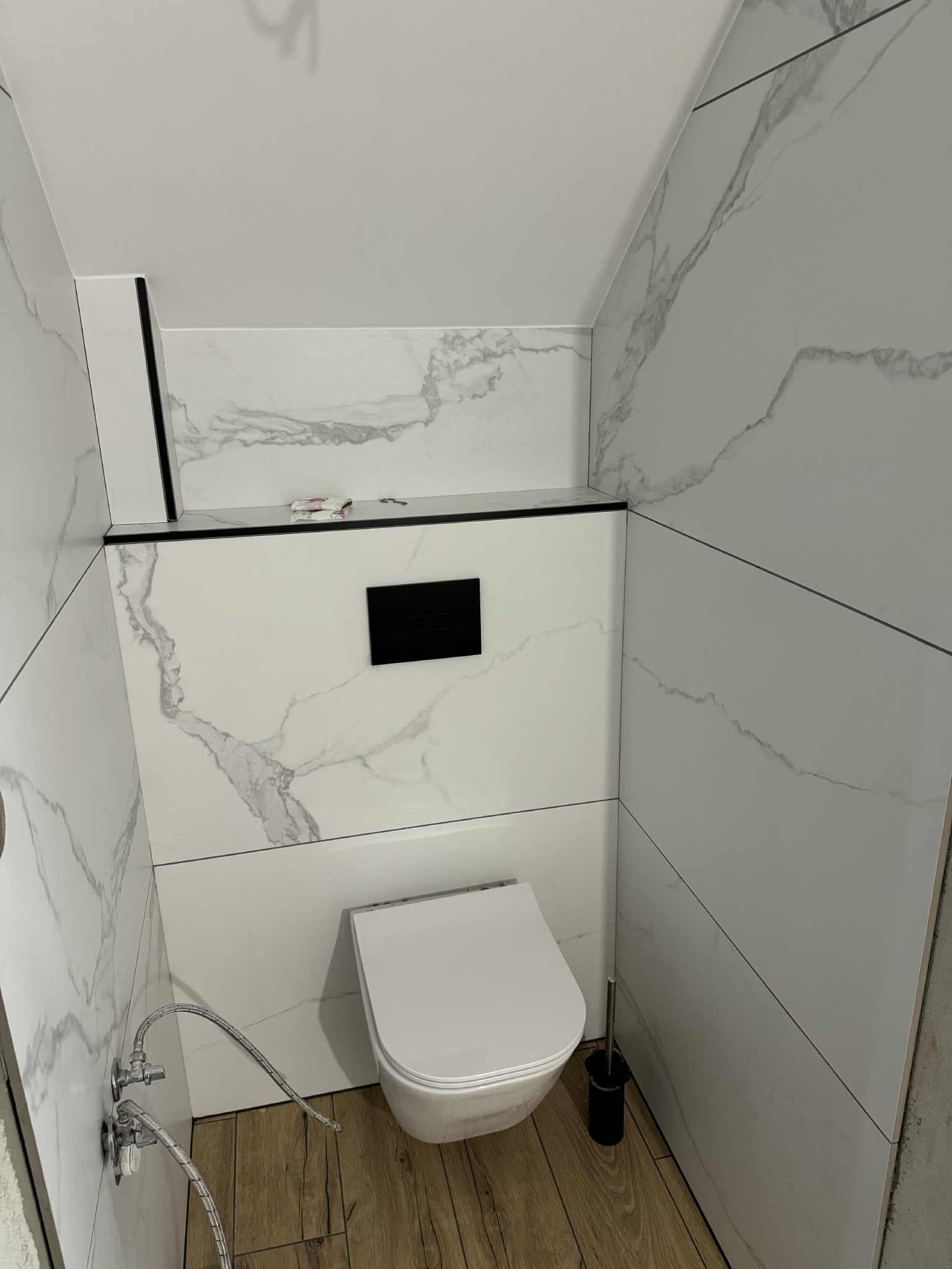 Nowoczesna toaleta w łazience wykończonej białymi płytkami imitującymi marmur, z drewnianą podłogą. Widoczne zawory i szczotka do WC.
