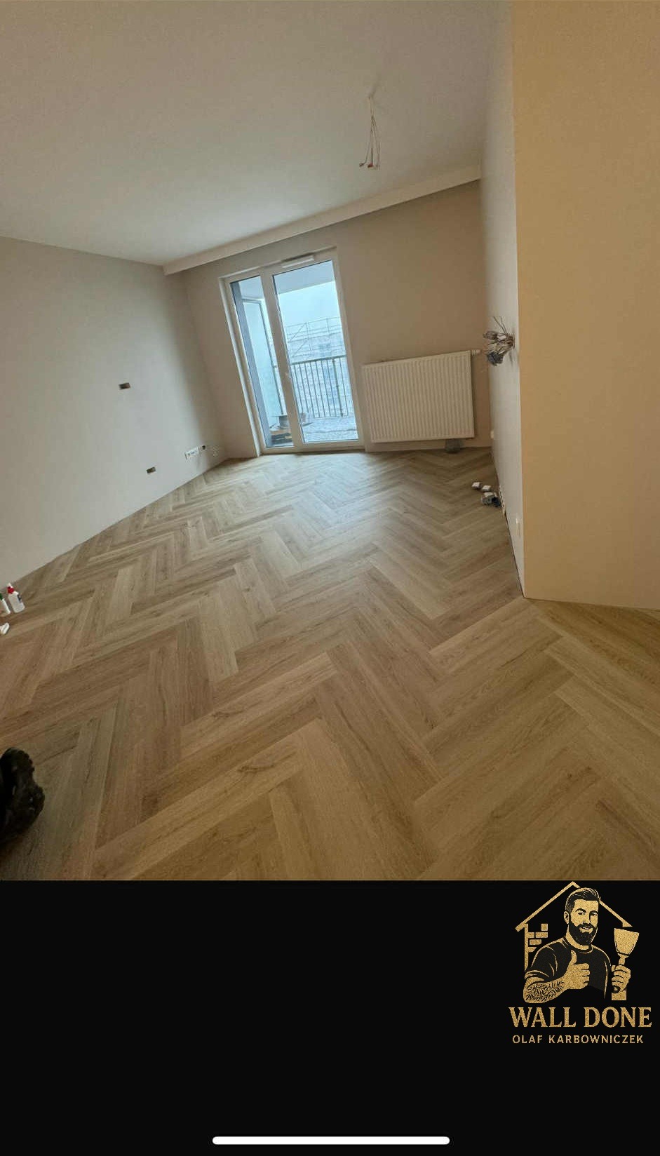 Jasne, parkietowe panele ułożone w jodełkę w pustym pokoju z beżowymi ścianami. Widoczne okno balkonowe i grzejnik. Minimalistyczne wykończenie wnętrza.