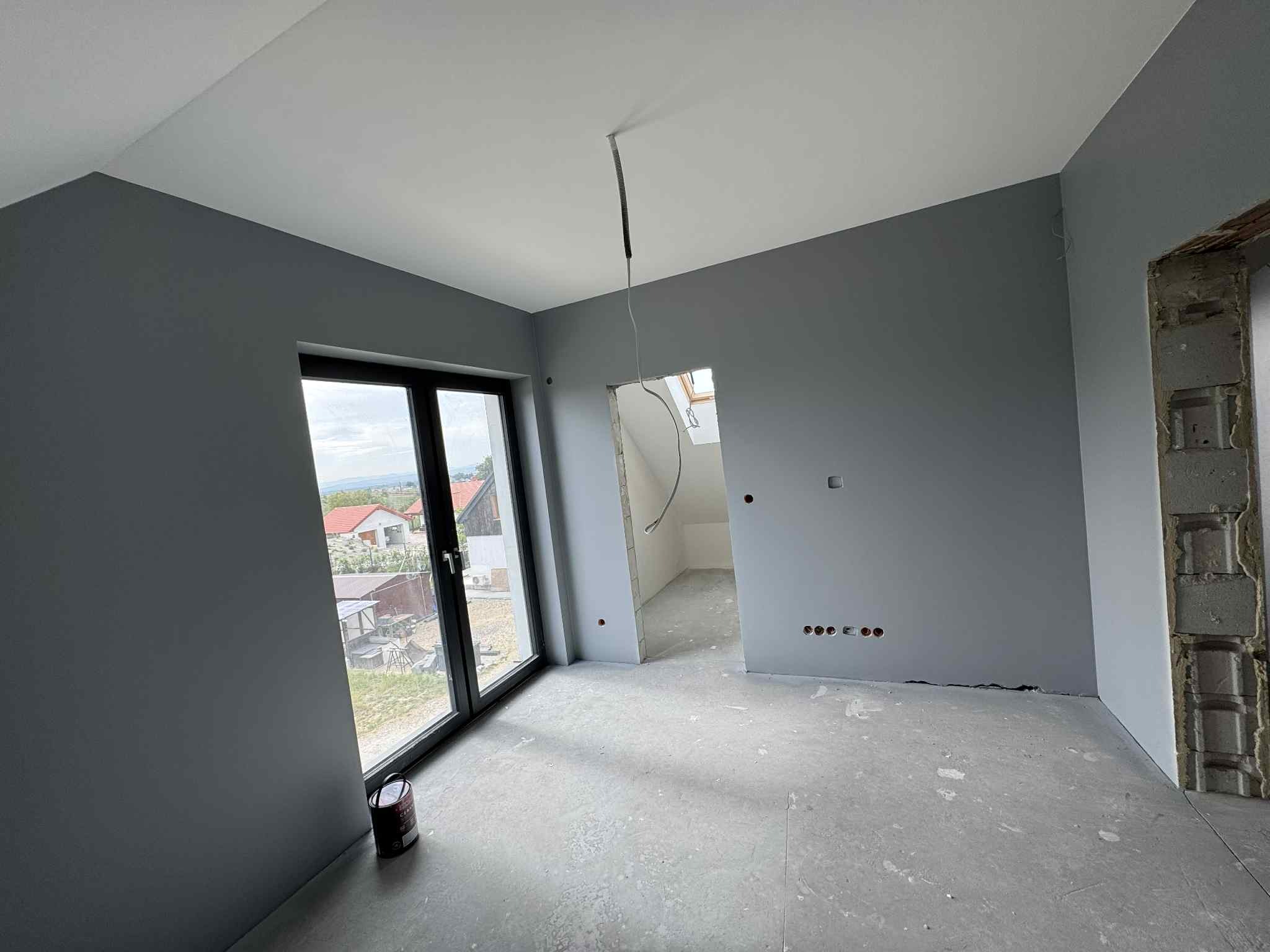 Pomieszczenie po malowaniu na szaro, widoczne okno z widokiem na domy, puszka farby przy ścianie, surowy stan wykończenia przy otworze drzwiowym.