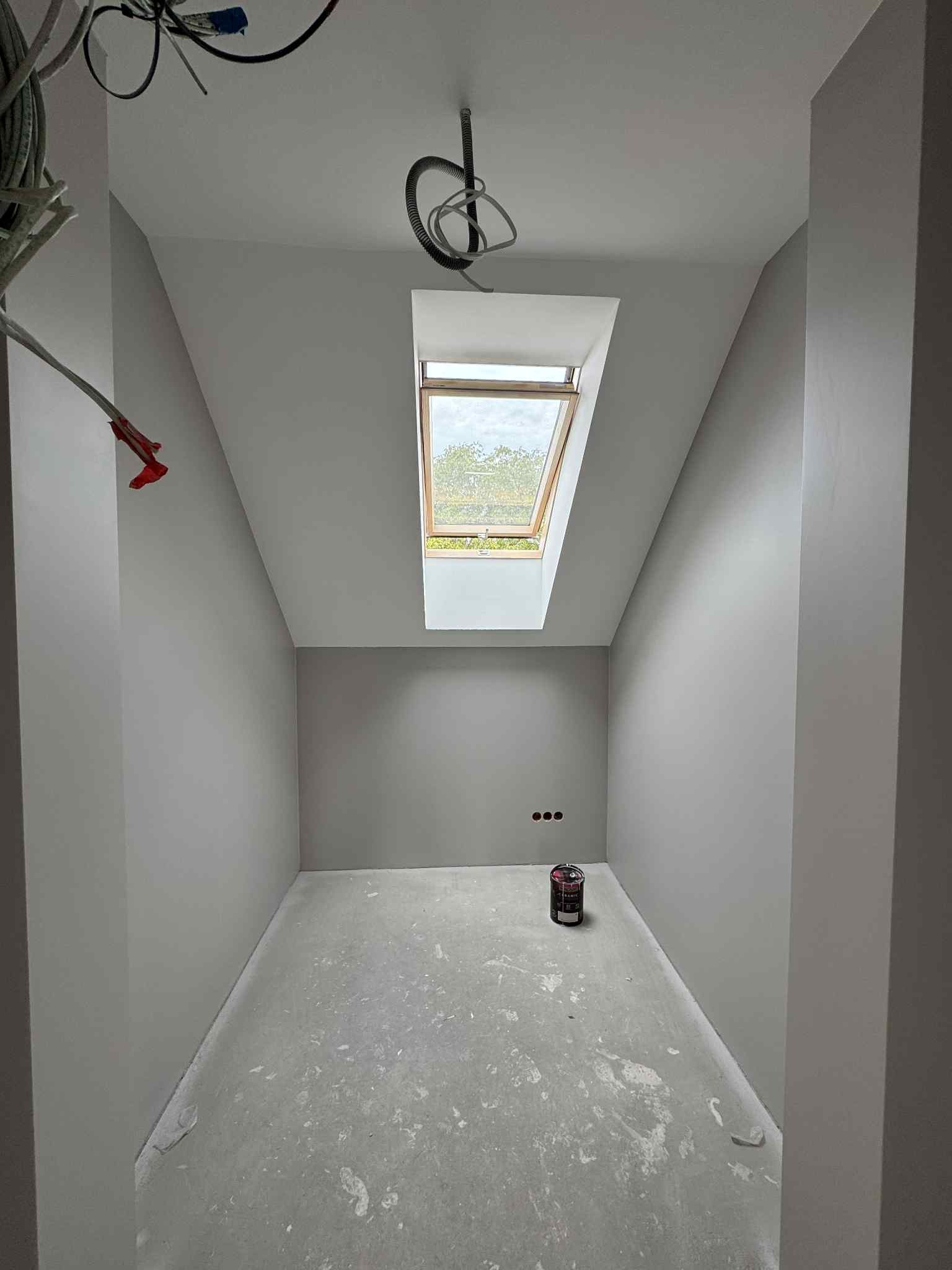 Pokój na poddaszu po szpachlowaniu i malowaniu na szaro, widoczne okno dachowe, puszki elektryczne i wiadro z farbą. Podłoga betonowa, przygotowana do dalszych prac.