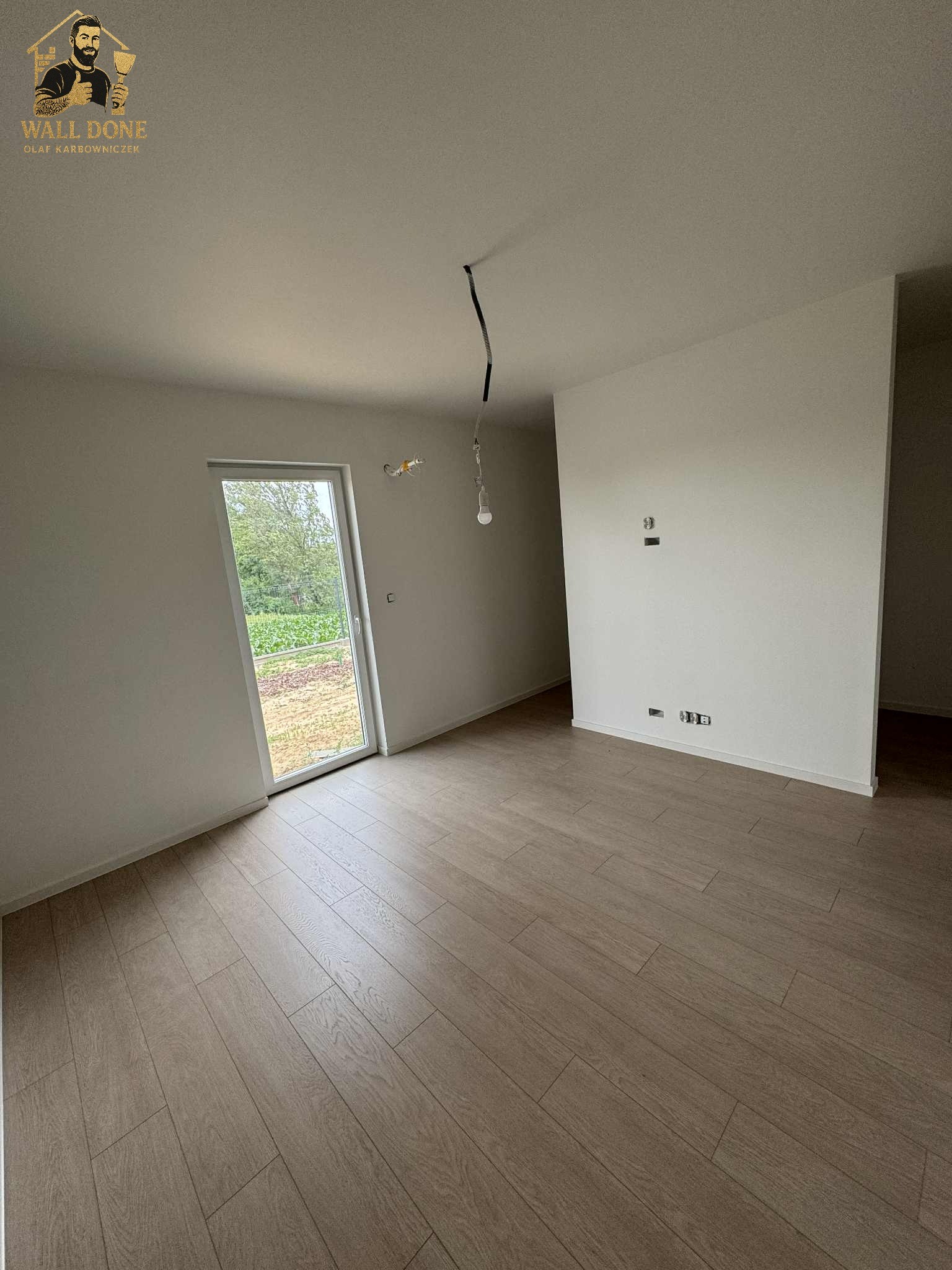 Jasne, puste wnętrze po remoncie z drewnopodobną podłogą, białymi ścianami i oknem z widokiem na zieleń. Widoczne gniazdka elektryczne i przewody.