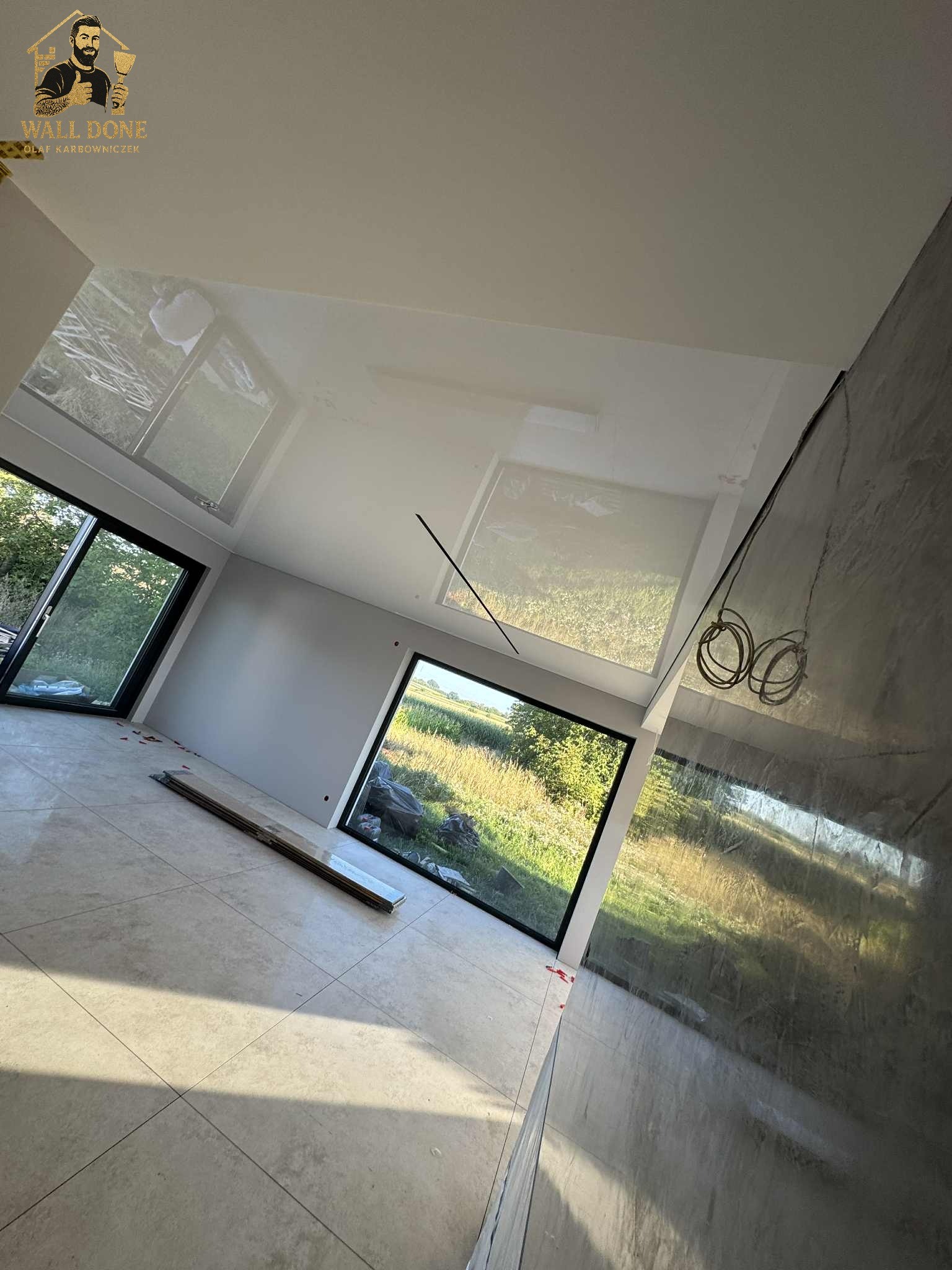 Wnętrze po pracach wykończeniowych: błyszczący sufit odbija widok z okna, szara ściana z efektem dekoracyjnym, jasna podłoga z płytek. Widoczne okna z widokiem na zewnątrz.
