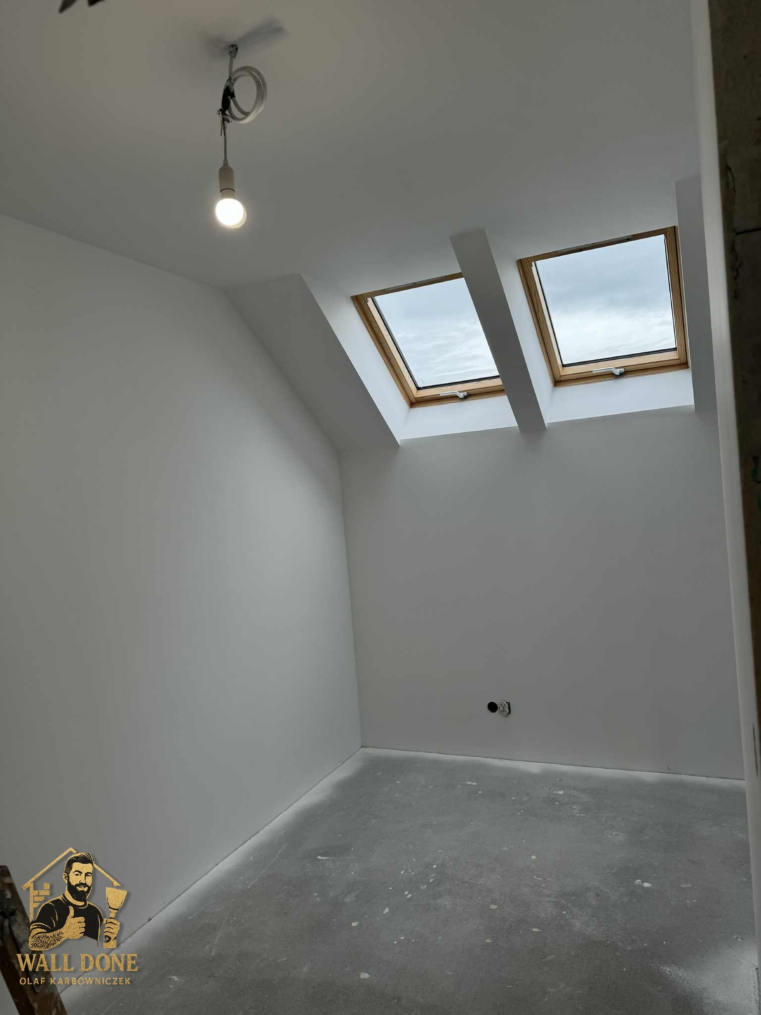 Jasne poddasze po remoncie z dwoma oknami dachowymi i szarą podłogą. Widoczna żarówka na kablu i logo firmy w rogu. Ściany świeżo pomalowane na biało.