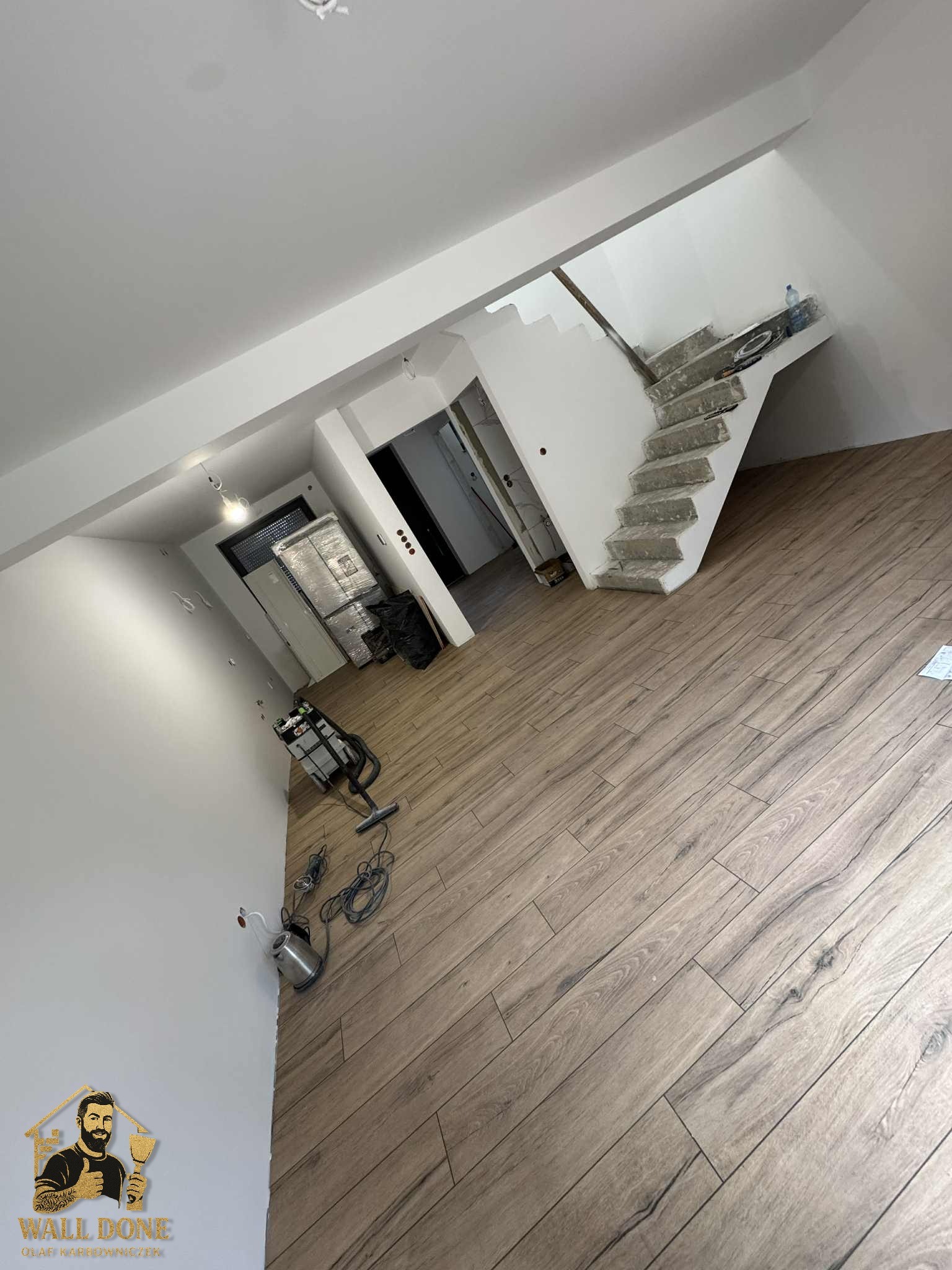 Wnętrze w trakcie prac wykończeniowych w Proszowicach: drewnopodobna podłoga, wyszpachlowane ściany i schody, narzędzia pracy. Widok na otwarte pomieszczenie.