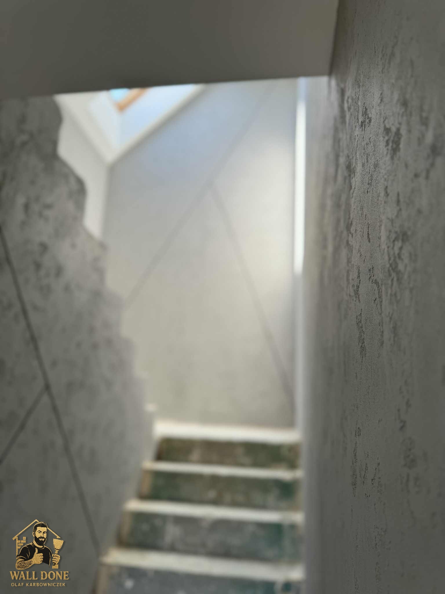 Wykończona klatka schodowa z dekoracyjną szarą ścianą o wyraźnej fakturze. Widoczne schody i górne okno. Na dole logo firmy.