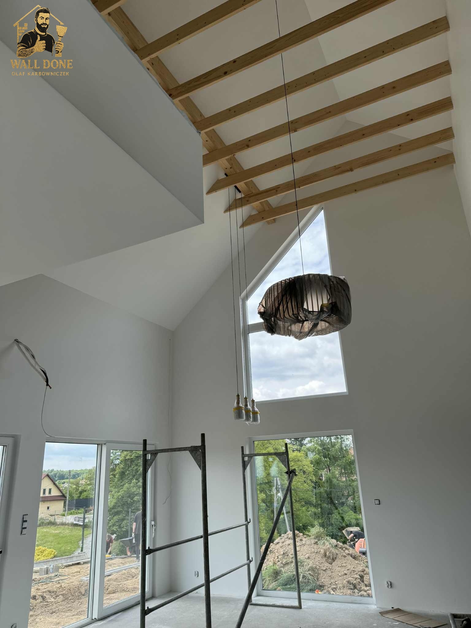 Wysokie wnętrze w trakcie wykańczania: białe ściany, drewniana konstrukcja dachu, okno trójkątne, wisząca lampa zabezpieczona folią, rusztowanie przy oknie.