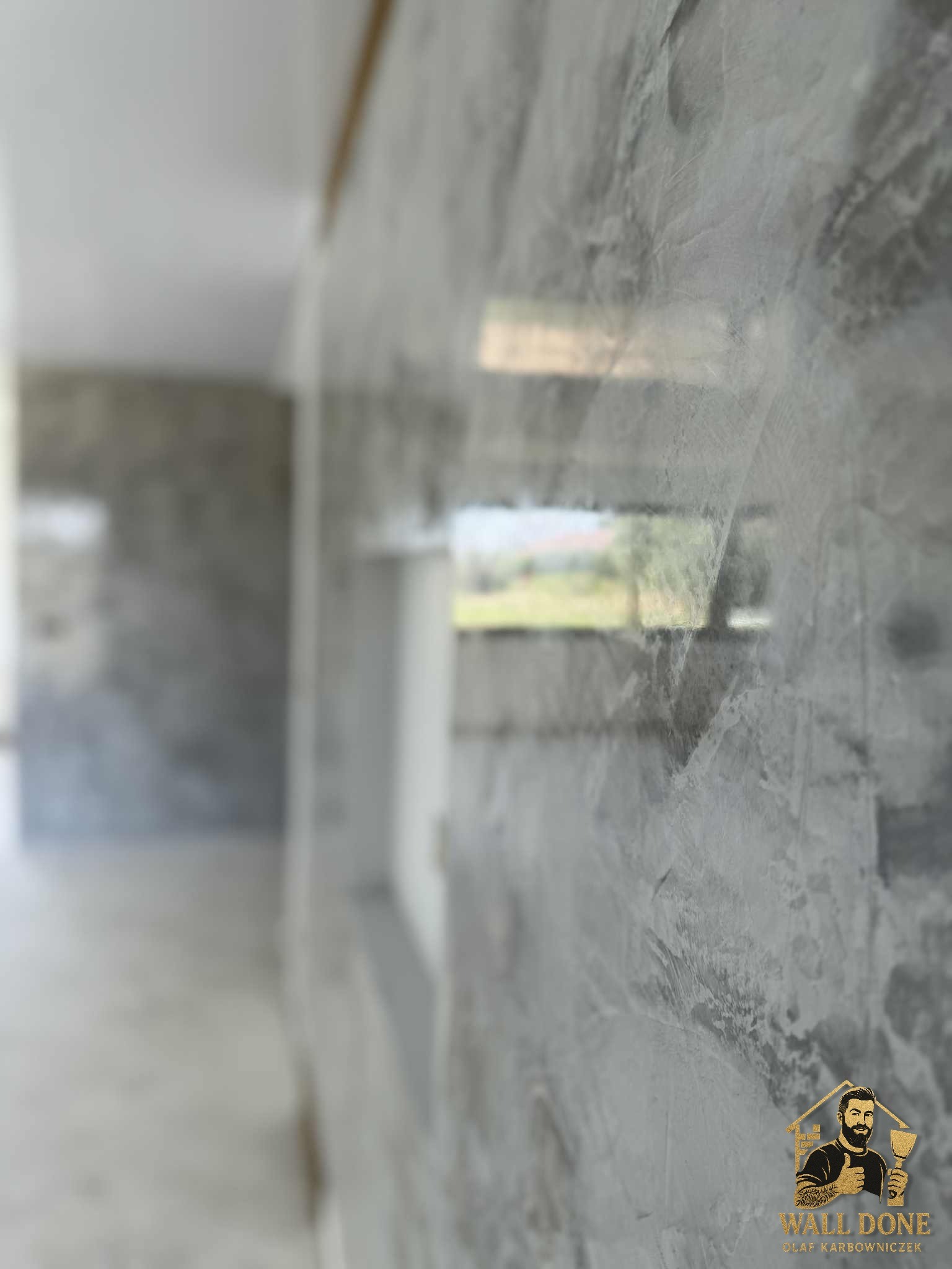 Ściana z efektem dekoracyjnym w odcieniach szarości, z widocznym refleksem światła i logo firmy w prawym dolnym rogu. Widok z perspektywy, z rozmytym tłem.