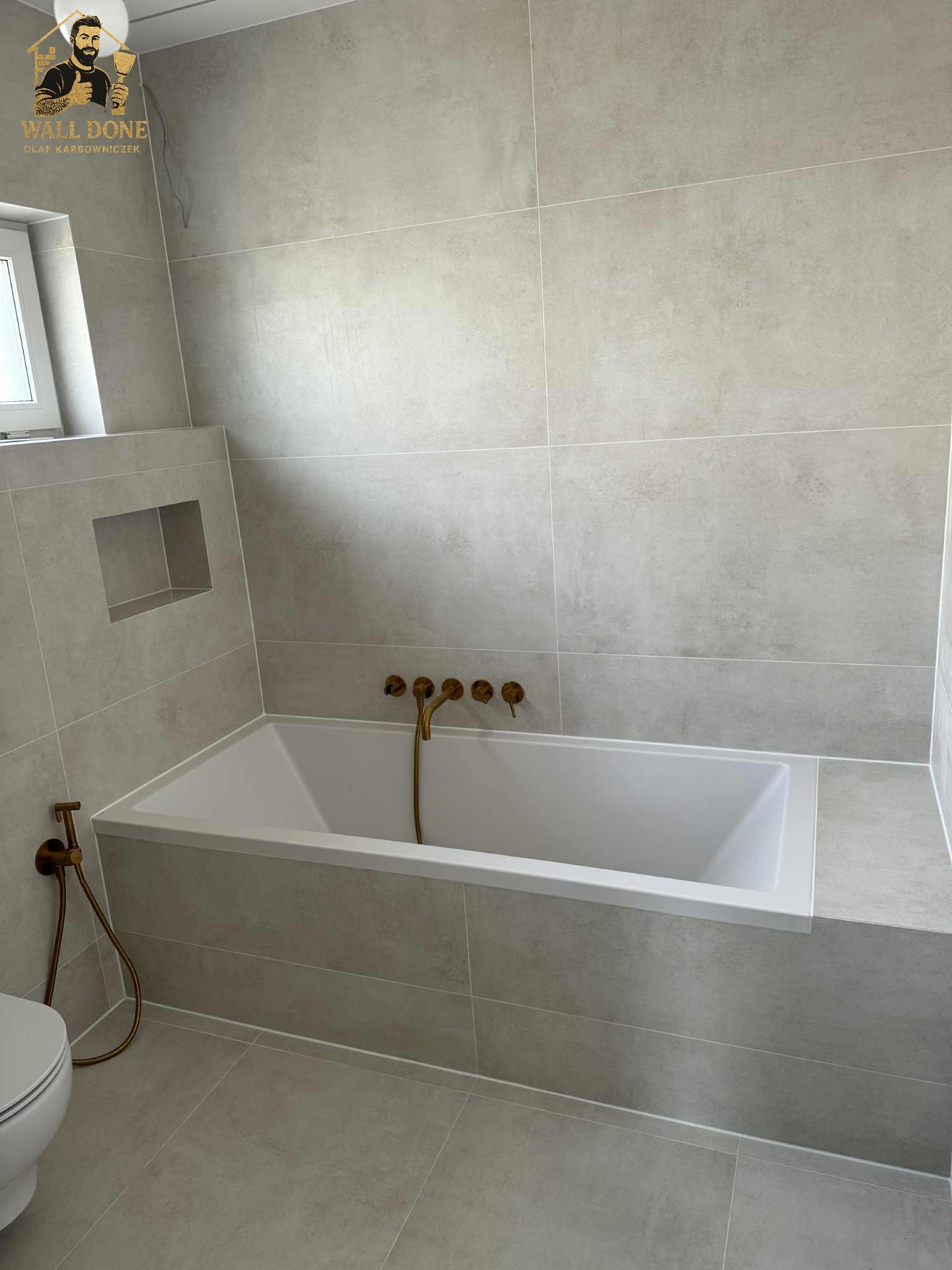 Wykończona łazienka z wanną, szarą glazurą i złotą armaturą. Widoczna wnęka w ścianie. Minimalistyczny design z dbałością o detale.