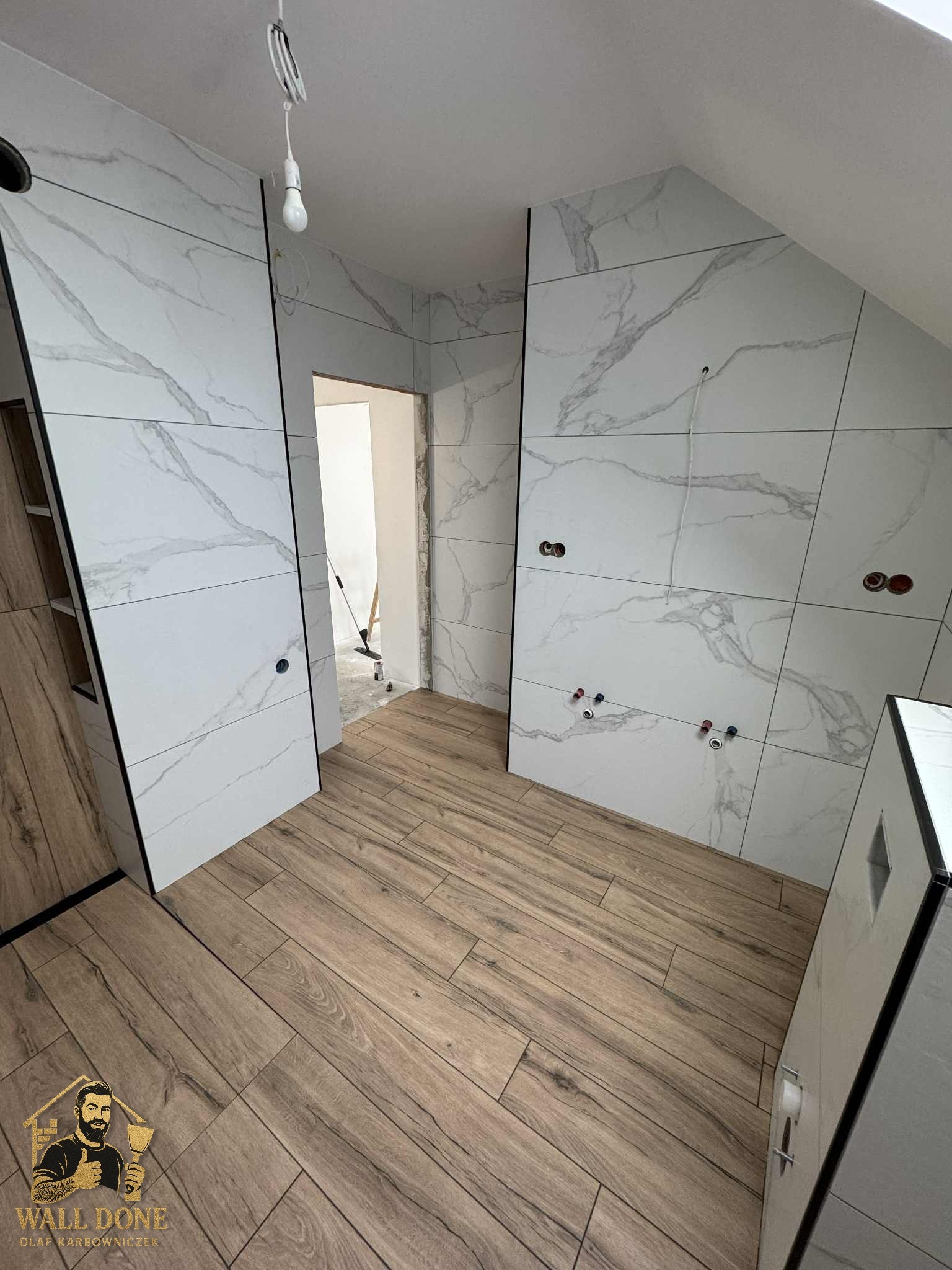 Wykończona łazienka na poddaszu z drewnopodobną podłogą i marmurowymi płytkami ściennymi z czarnymi listwami. Widoczne punkty instalacyjne.