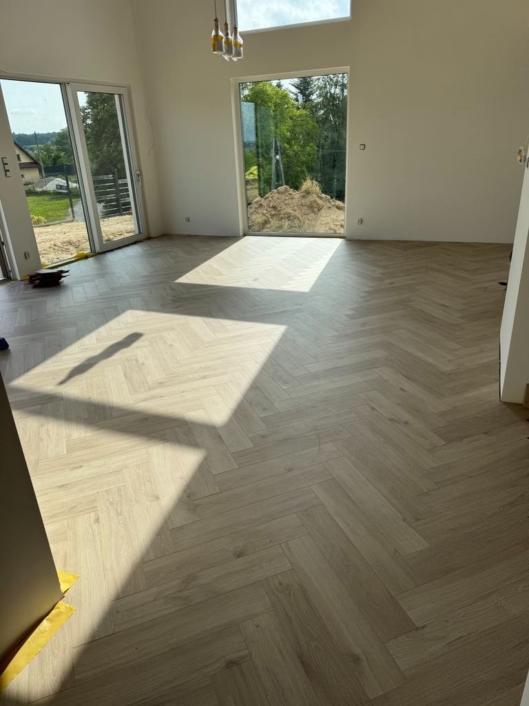 Jasna, drewniana podłoga ułożona w jodełkę francuską w przestronnym, jasnym pokoju z dużymi oknami. Widoczne promienie słoneczne padające na podłogę.