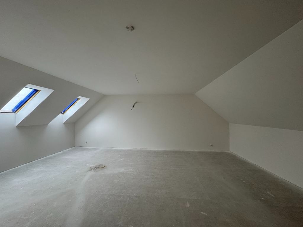 Poddasze po pracach wykończeniowych: białe ściany, skośny sufit z dwoma oknami dachowymi. Podłoga szara, surowa. Widoczne przewody elektryczne.