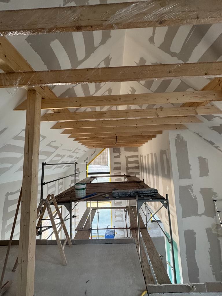 Skośny sufit poddasza w trakcie prac wykończeniowych. Widoczne rusztowanie, drewniane belki i ściany pokryte wstępną warstwą gładzi. Okno w tle.