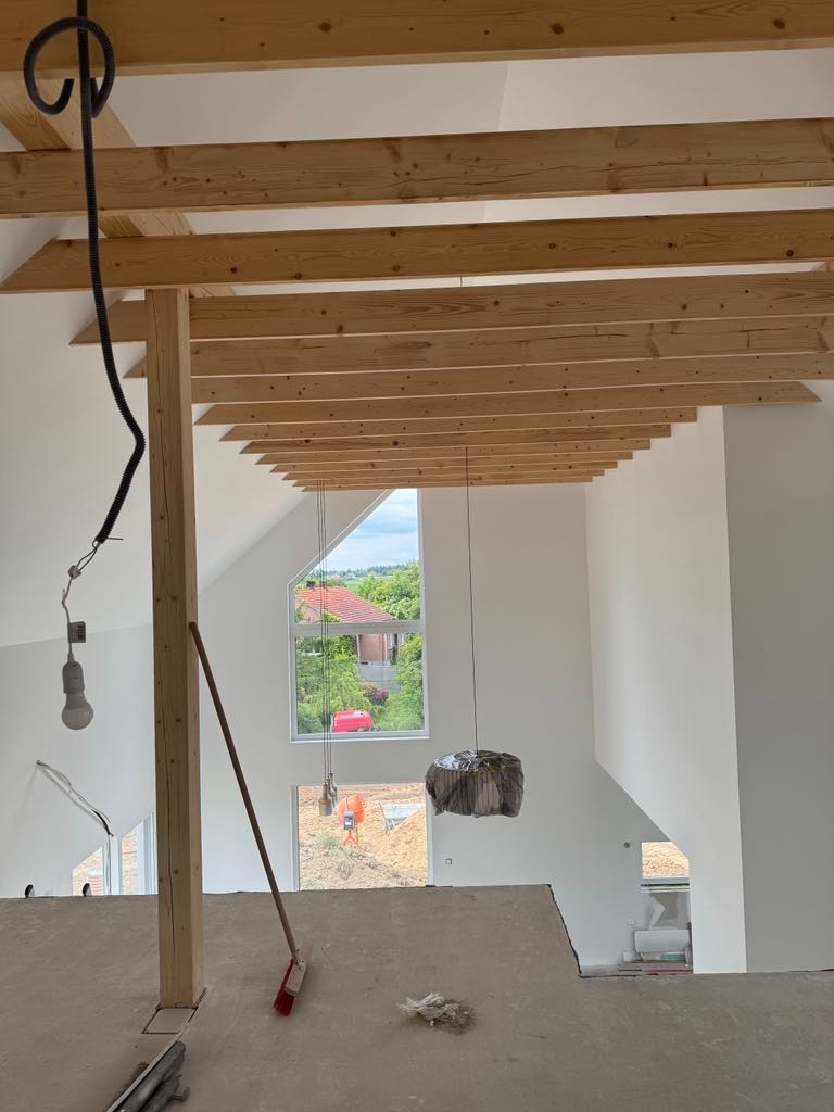 Surowe drewno stropu w jasnym wnętrzu z oknami. Widoczne narzędzia i niedokończone prace. Elementy instalacji elektrycznej zwisają z sufitu. W tle widok na zewnątrz.