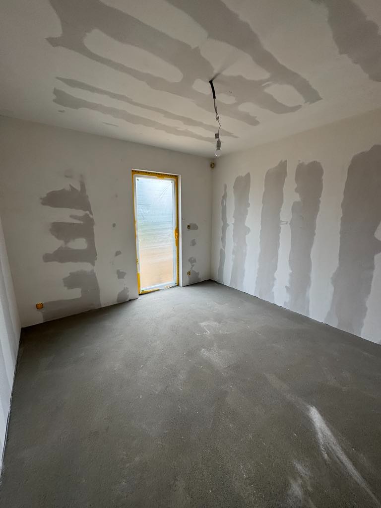 Pokój w trakcie prac wykończeniowych: szpachlowane ściany z widocznymi pasami, wylewka podłogowa, okno zabezpieczone folią, surowa żarówka.
