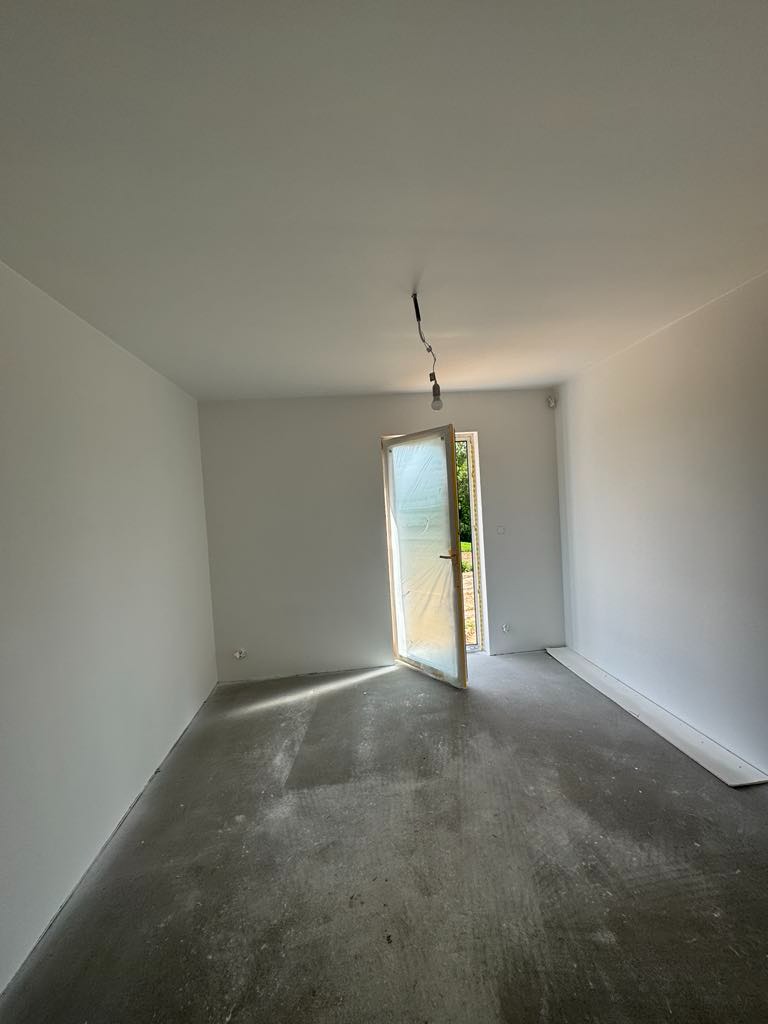 Jasne wnętrze po remoncie: białe ściany, surowa podłoga, otwarte drzwi z folią ochronną. Widok na zieleń za oknem. Minimalistyczny wystrój, surowe wykończenie.