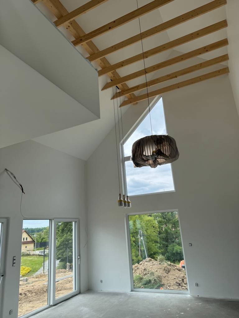 Wysokie wnętrze w stanie deweloperskim z drewnianą konstrukcją sufitu, dużymi oknami i wiszącą lampą w folii ochronnej. Świeżo pomalowane ściany na biało.