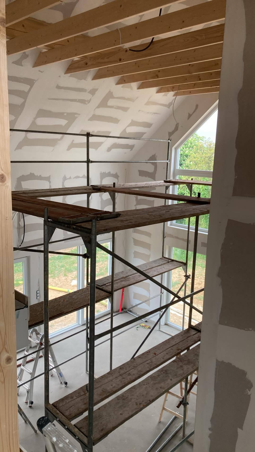 Wnętrze w trakcie prac wykończeniowych: rusztowanie, ściany z nałożoną gładzią, drewniana konstrukcja dachu. Widok z góry na pomieszczenie z oknami.