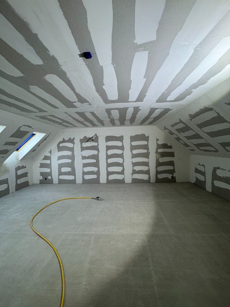 Poddasze w trakcie prac wykończeniowych: płyty gipsowe na skosach, częściowo zaszpachlowane łączenia, żółty wąż na szarej podłodze, okno dachowe.