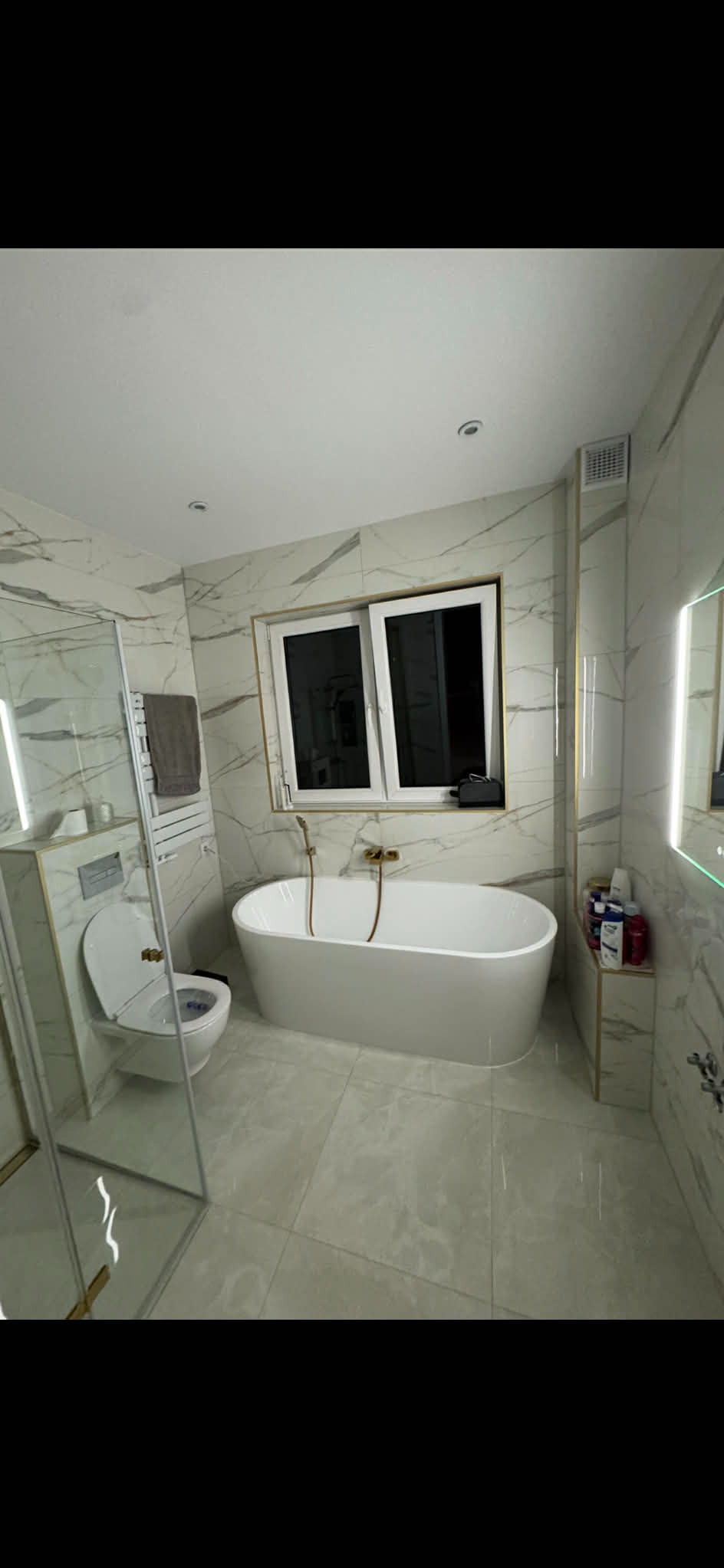 Elegancka łazienka z wanną wolnostojącą, marmurowymi płytkami i złotymi akcentami. Widoczne okno i kabina prysznicowa ze szklanymi drzwiami.