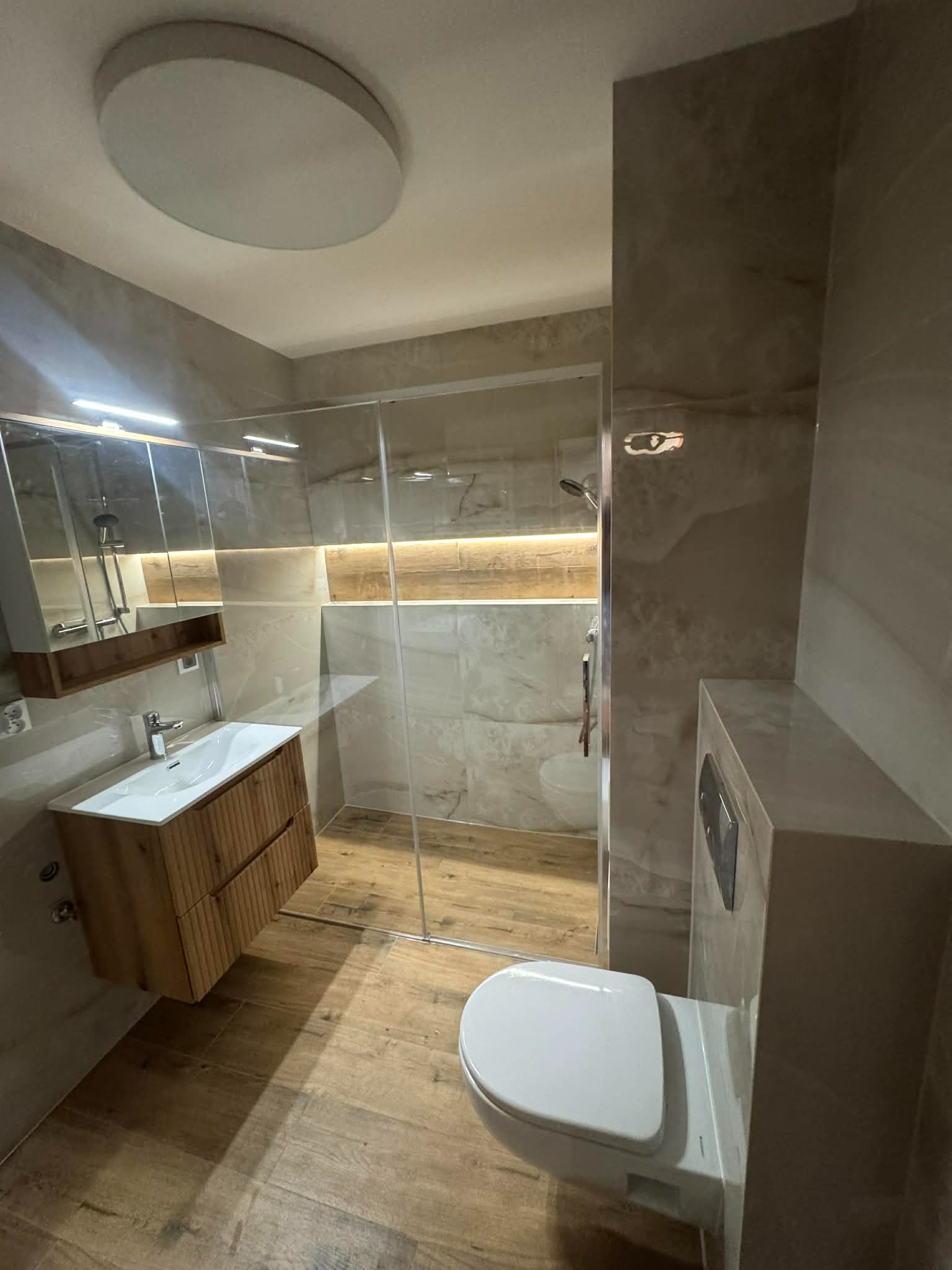 Nowoczesna łazienka z kabiną prysznicową typu walk-in, drewnianą szafką pod umywalką i beżowymi płytkami imitującymi marmur. Oświetlenie LED podkreśla detale.