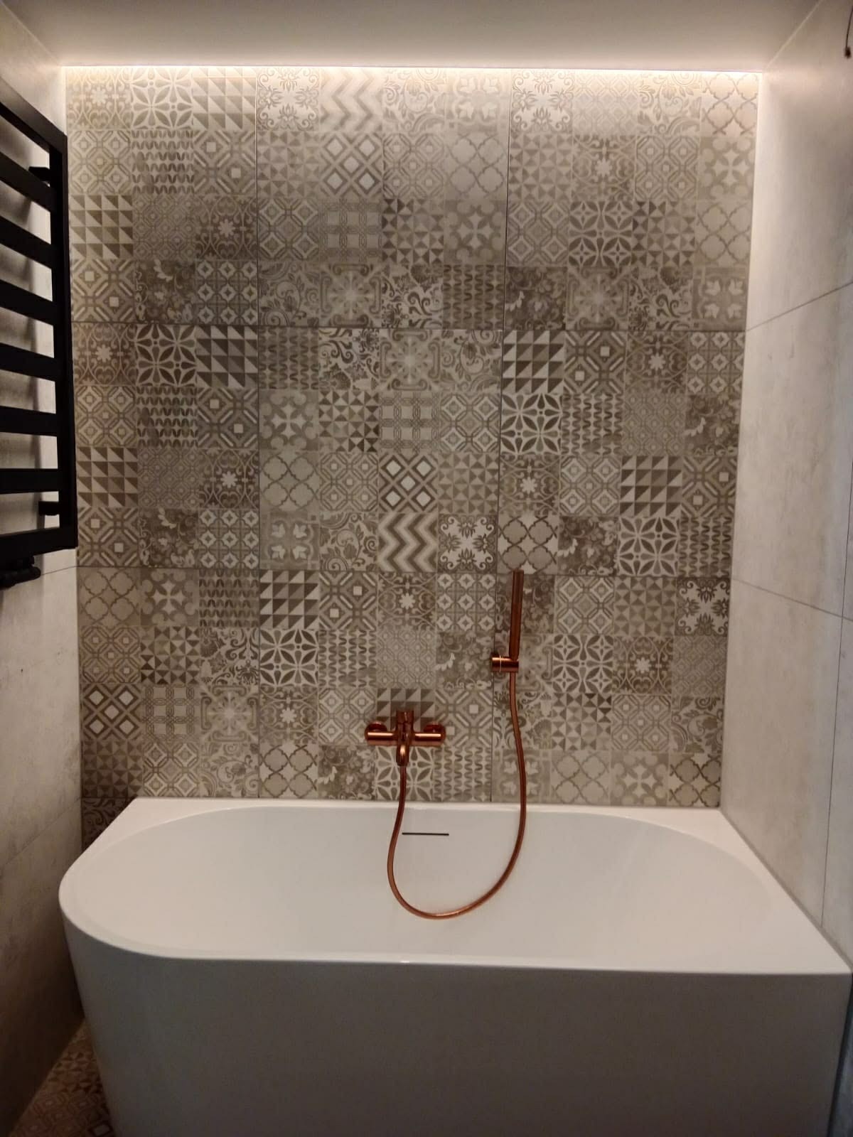 Wykończona łazienka z wanną, mozaiką na ścianie i miedzianą armaturą. Podświetlenie LED nadaje wnętrzu nowoczesny charakter. Grzejnik drabinkowy po lewej stronie.