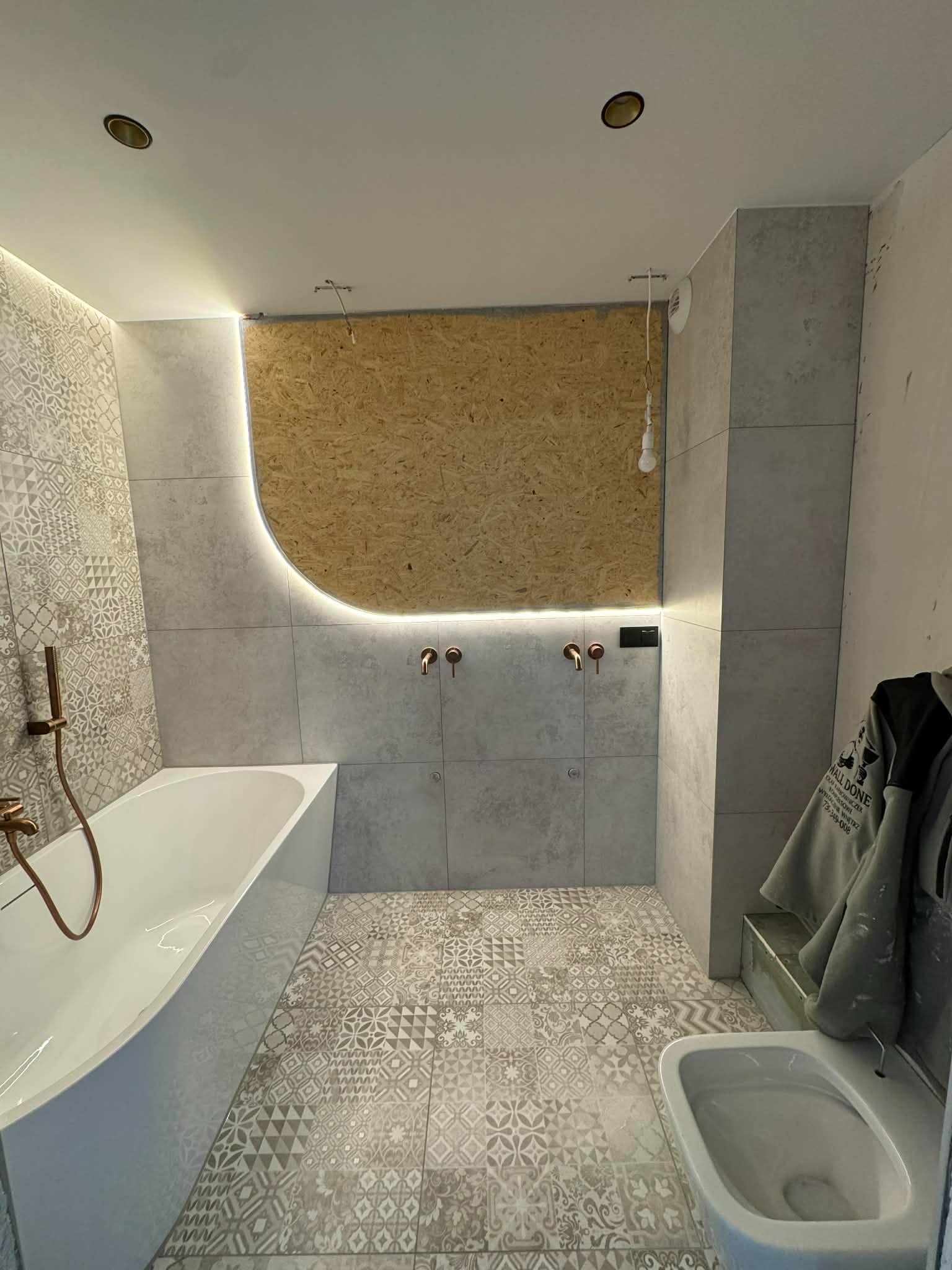 Wykończona łazienka z mozaikową podłogą i ścianą, podświetlona LED. Widoczna wanna, toaleta i fragment ściany z surową płytą OSB. Armatura w kolorze miedzi.