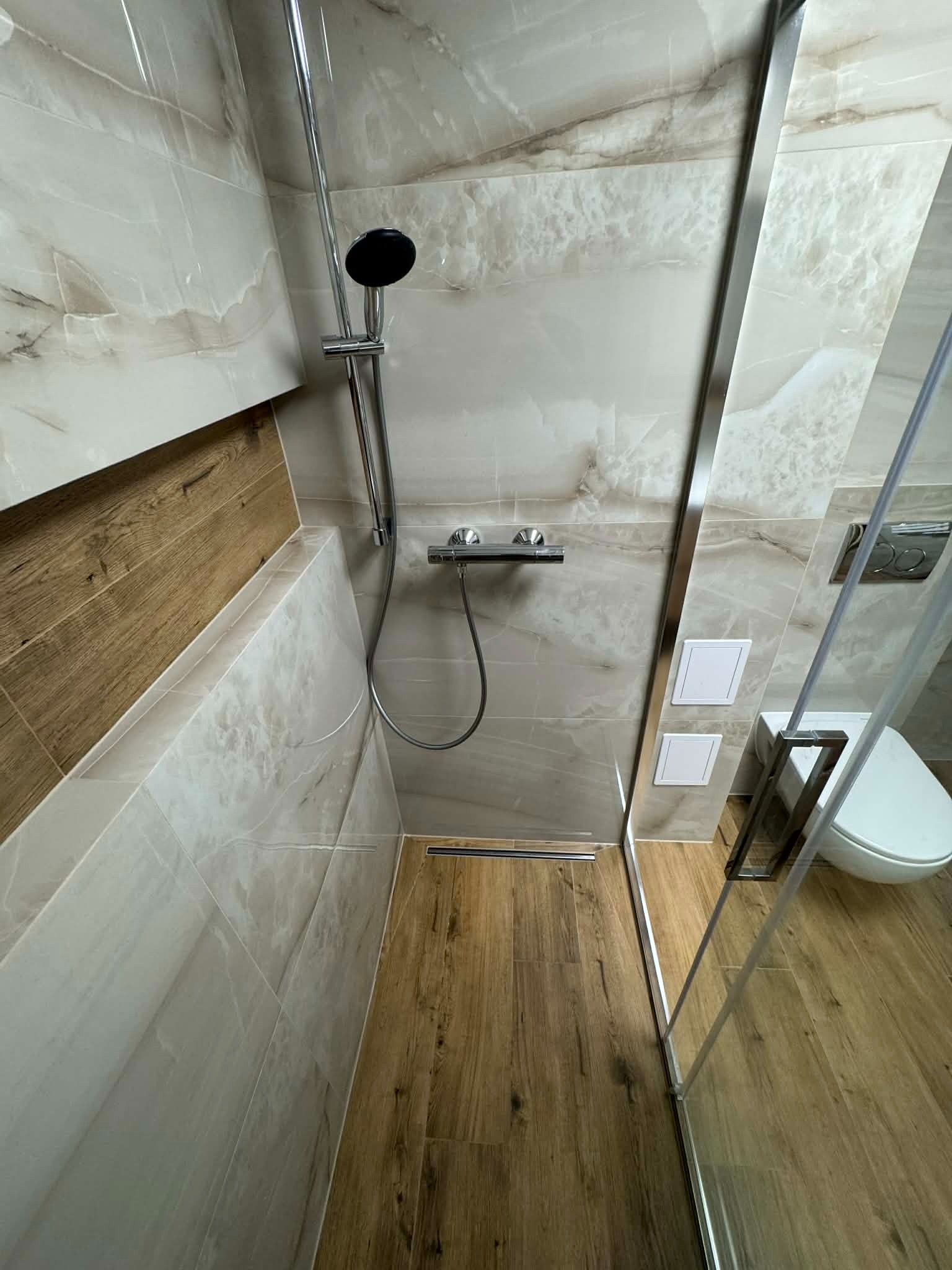 Nowoczesna kabina prysznicowa typu walk-in z drewnopodobną podłogą i marmurowymi płytkami ściennymi, widoczny odpływ liniowy i toaleta w tle.