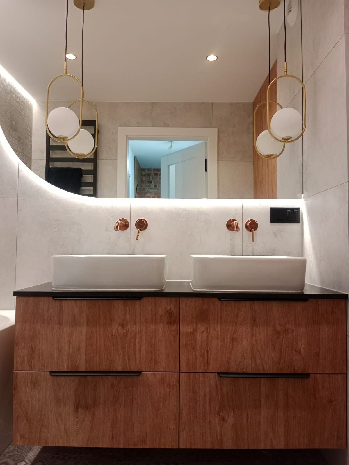 Nowoczesna łazienka z podwójną umywalką nablatową na drewnianej szafce, oświetlona wiszącymi lampami z elementami złota i lustrem z podświetleniem LED.